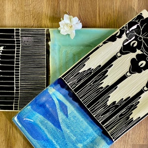 May include: Three rectangular ceramic serving trays with black and white designs. The first tray has a black background with white geometric patterns. The second tray has a light blue background with white and black abstract designs. The third tray has a white background with black floral designs.
