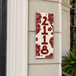 May include: A rectangular ceramic house number plaque featuring the digits 2118 in a deep red glaze. The plaque has a cream-colored background decorated with embossed red floral patterns along the vertical edges and two small mounting holes.