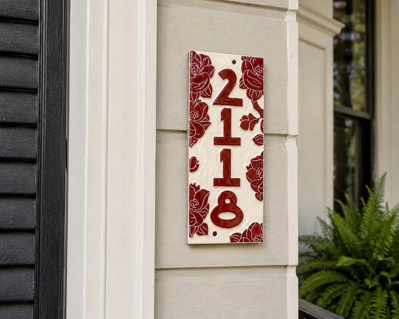 May include: A rectangular ceramic house number plaque featuring the digits 2118 in a deep red glaze. The plaque has a cream-colored background decorated with embossed red floral patterns along the vertical edges and two small mounting holes.