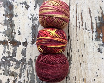 May include: Three balls of yarn stacked on top of each other. The top ball is yellow and red, the middle ball is red and yellow, and the bottom ball is a deep red.