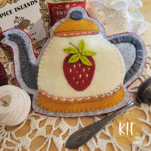 May include: A felt teapot-shaped kit with a strawberry applique. The teapot is cream-colored with a gray handle and spout, and a yellow lid. The strawberry is red with green leaves. The kit includes a tea infuser and a ball of white thread.