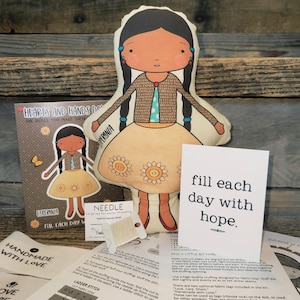 May include: A fabric doll kit featuring a printed doll named Esperanza with black braided hair, a brown cardigan, and a tan skirt with flower patterns. The kit includes an instruction sheet, a needle, thread, and a card with the text fill each day with hope.