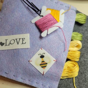 May include: A lavender felt sewing kit with spools of pink, yellow, and green thread. A small patch with a bee illustration is stitched on the front, along with a label that says "LOVE". A black paperclip holds the thread spools.