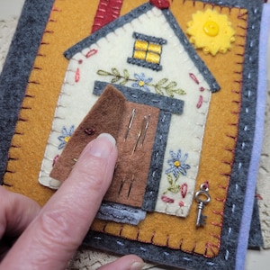 May include: A handmade felt house-shaped needle case. The case features a cream-colored house with a brown door, a yellow window, and a red chimney. The door opens to reveal needles. A yellow button sun and a silver key are also included.
