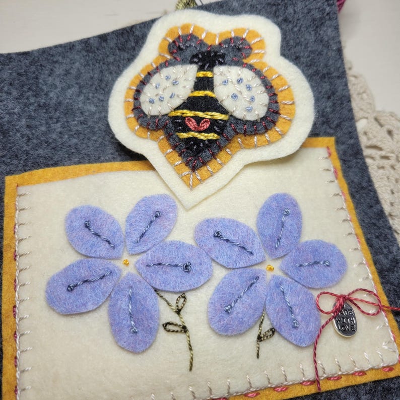 May include: A handmade felt art piece featuring a bee and flowers. The bee is yellow, black, and gray, with beaded accents. The flowers are lavender with silver thread details. The piece is on a gray background with yellow and red stitching.