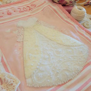 May include: A handmade, off-white wedding dress with intricate floral embroidery and lace sleeves is displayed on a pink and white striped fabric. The date "6.2025" is embroidered in red thread. A delicate, crocheted piece is visible.