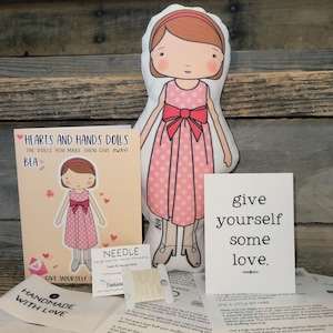 May include: A fabric doll kit featuring a printed doll named Bea in a pink polka dot dress with a red bow, an instruction card titled Hearts and Hands Dolls, a needle, a spool of thread, and a card with the text give yourself some love.