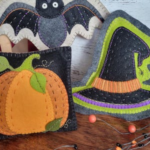 Pode incluir: Decorações de feltro de Halloween feitas à mão, incluindo um morcego, uma abóbora e um chapéu de bruxa. O morcego é cinza e preto com detalhes roxos. A abóbora é laranja com folhas verdes. O chapéu de bruxa é preto, verde e laranja.