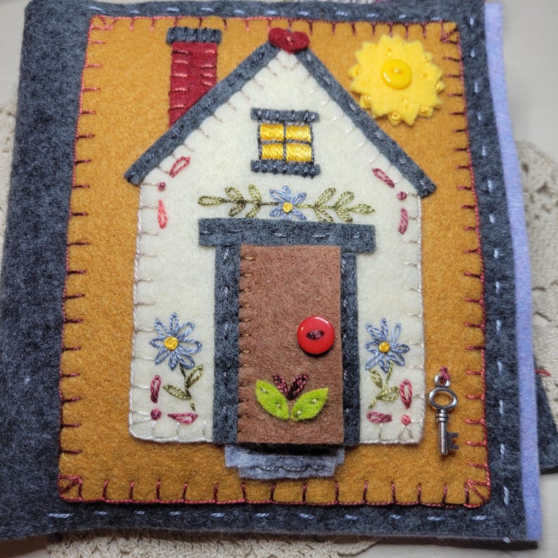 May include: Handmade felt book cover featuring a house design. The cover has a white house with a brown door, red chimney, and yellow window. The house is on a mustard-colored background, with gray borders and a key charm.
