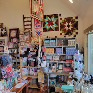 May include: A well-lit shop interior with wooden shelves and tables displaying various handcrafted items. Quilts with geometric patterns hang on the walls. The shop offers a variety of products, including cards, mugs, and decorative objects.