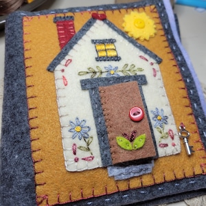 May include: A handmade felt house-shaped item with a cream-colored roof, brown door, and yellow background. The house features a red chimney, a yellow sun, and embroidered blue flowers. A small key is visible.