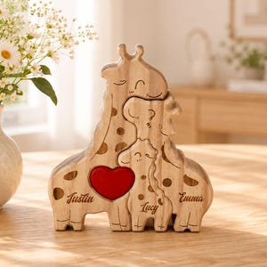Op de afbeelding: Een houten giraffenfamilie puzzelfiguur bestaande uit drie in elkaar grijpende giraffenvormen met natuurlijke houtnerf, donkerbruine vlekken en een felrood hart. De namen Justin, Lucy en Emma zijn in het hout gegraveerd.