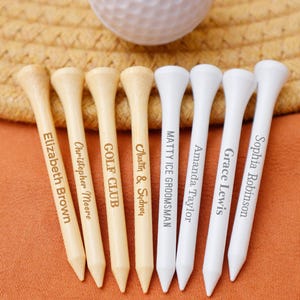 Peut inclure: Un ensemble de tees de golf personnalisés, certains avec des noms et d'autres avec du texte comme "GOLF CLUB". Les tees sont en bois et en plastique blanc, disposés sur une surface orange avec une balle de golf en arrière-plan.