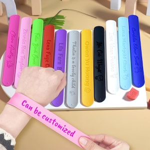 May include: A collection of colorful, personalized wristbands in various shades, including pink, purple, green, and yellow. Each band features a name and a small graphic. One pink band in the foreground reads "Can be customized."