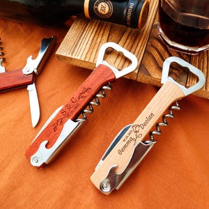 May include: Two personalized wine bottle openers with wooden handles and stainless steel components. One is reddish-brown with "S C" and a date engraved. The other is light wood with "Gemma Declan" and a date. A wine bottle and glass are in the background.