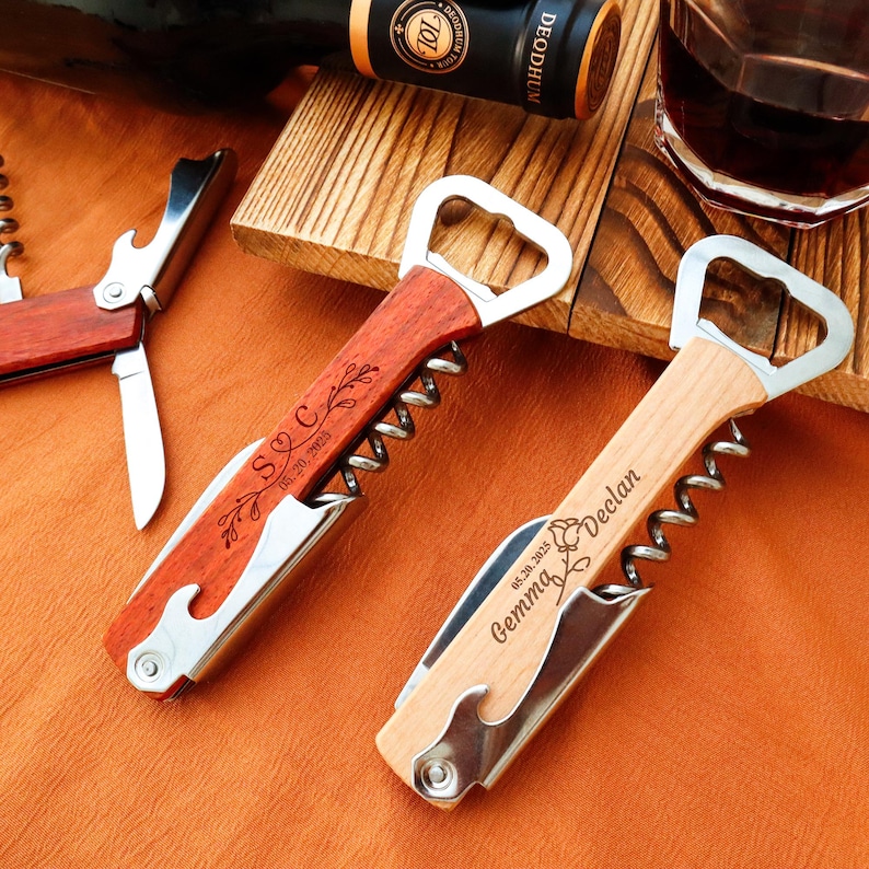 May include: Two personalized wine bottle openers with wooden handles and stainless steel components. One is reddish-brown with "S C" and a date engraved. The other is light wood with "Gemma Declan" and a date. A wine bottle and glass are in the background.