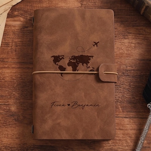 Può includere: Un diario in similpelle marrone con una mappa del mondo marrone scuro, una rotta di volo tratteggiata e un piccolo aereo. La copertina è personalizzata con i nomi Fiona e Benjamin separati da un cuore e fissata da un elastico.