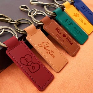 Peut inclure: Plusieurs porte-clés personnalisés en cuir de différentes couleurs : rouge, beige, marron, vert, jaune et bleu. Chaque porte-clés est doté d'un anneau métallique et gravé de noms, d'initiales ou de dates. Ils sont disposés sur une surface texturée.