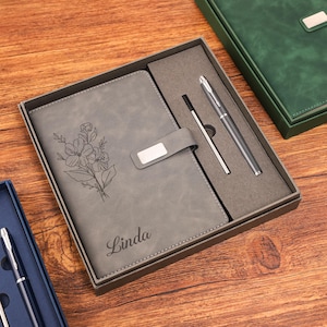 Op de afbeelding: Een grijze notitieboek- en pennenset in een geschenkdoos. Het notitieboek heeft een bloemenontwerp en de naam "Linda". De set bevat een zilveren pen en een zwarte penvulling. De doos is grijs met een donkergrijs interieur.