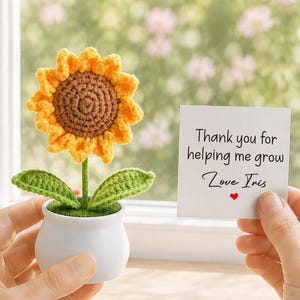 Può includere: Un girasole all'uncinetto con petali gialli e centro marrone, inserito in un piccolo vaso bianco con foglie verdi. Accanto viene tenuto un cartoncino quadrato bianco con il testo stampato: Thank you for helping me grow, Love Iris, e un piccolo cuore rosso.