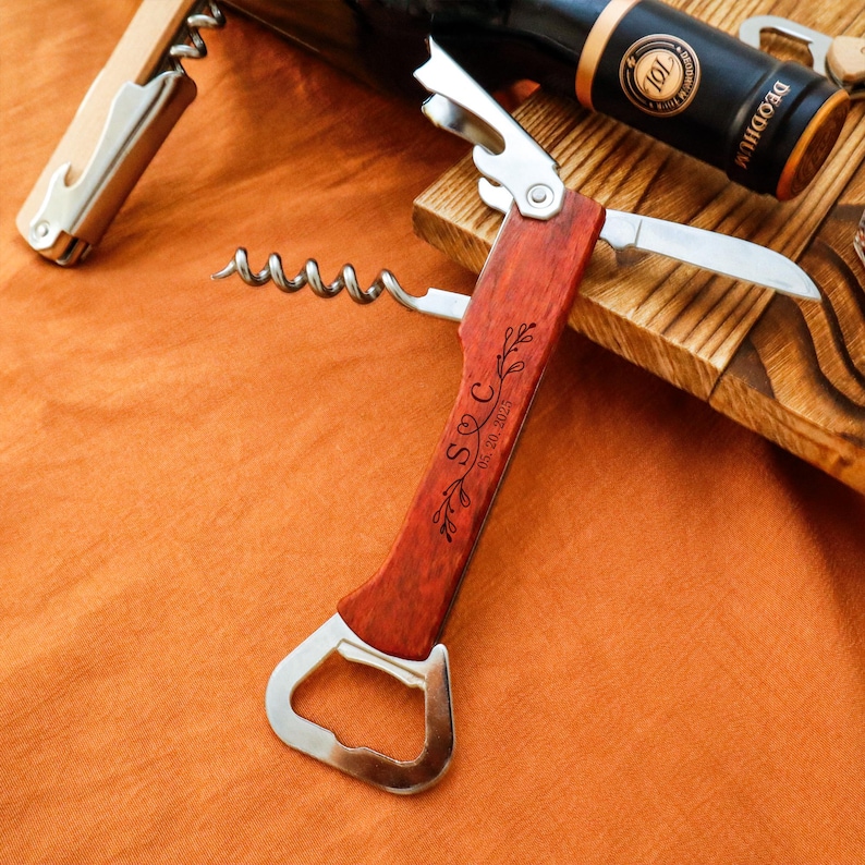 May include: A wooden-handled wine opener with a corkscrew, bottle opener, and knife. The handle is engraved with initials and a date. A bottle of wine and another corkscrew are in the background. The opener is on an orange surface.