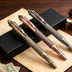 May include: Three metal pens with a twisted, textured grip and a smooth upper barrel featuring a clip and the name Arthur engraved on the side. The pens are finished in antique brass, copper, and gunmetal tones, each with a pointed tip and cap.