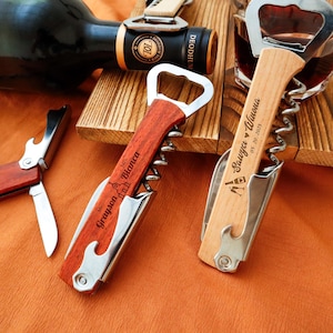 May include: Two personalized wooden wine bottle openers with silver accents. One is dark wood with "Mr. Grayson Bianca" engraved. The other is light wood with "Sawyer Winona 05.20.2025" engraved. A wine bottle and glass are in the background.