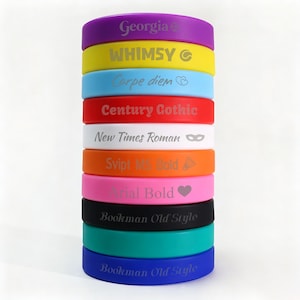 Pode incluir: Uma pilha vertical de dez pulseiras de silicone coloridas, cada uma com texto e símbolos gravados exclusivos. As pulseiras são roxa, amarela, azul clara, vermelha, branca, laranja, rosa, preta, azul-petróleo e azul, com várias fontes e ícones.