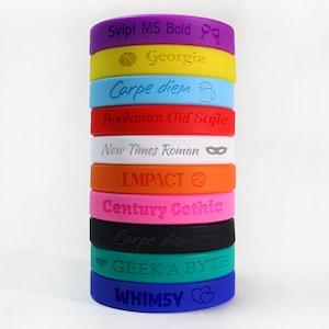 May include: A vertical stack of eleven colorful silicone wristbands featuring various debossed text and icons. The bands are purple, yellow, light blue, red, white, orange, pink, black, teal, and dark blue, displaying fonts like Script MS Bold.
