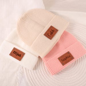 Puede incluir: Tres gorros de punto en crema, blanco y rosa. Cada gorro tiene un parche de cuero marrón con un nombre: "Elijah", "Caleb" y "Sophia". Los gorros están doblados en el borde.