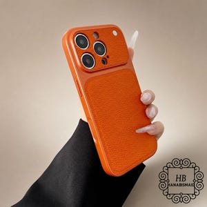 May include: A bright orange smartphone case with a textured, leather-like finish on the back panel and camera module area. The case features a raised camera cutout with three circular lens openings and a small circular flash opening at the top.
