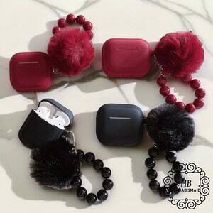 May include: AirPod cases in burgundy and black, some with fluffy pom-poms and beaded accents. One case is open, revealing white earbuds. The cases are on a marble surface, offering a stylish accessory.