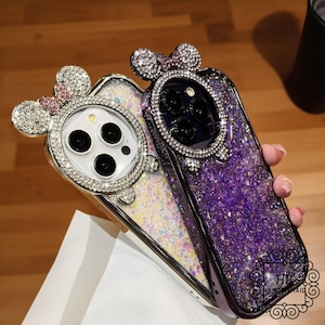 May include: Two sparkly phone cases with a glittery design. One case is purple with a black rhinestone Mickey Mouse head. The other case is white with a silver rhinestone Mickey Mouse head. Both cases have a glitter design.