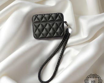 May include: Black quilted leather AirPod case with a braided wrist strap. The case has a diamond pattern and a silver-tone metal clasp. The background is a white satin fabric.