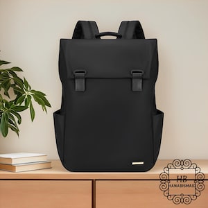 May include: A black backpack with a flap closure and two buckle straps. The backpack has two side pockets and a small gold-colored rectangular detail on the front. The bag is sitting on a wooden surface.
