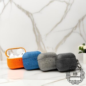 May include: Four fabric-covered AirPod cases in a row. The first is open, revealing white earbuds, and is orange. The others are blue, gray, and dark gray. The cases have a textured appearance and a small loop for a keychain.