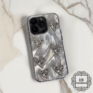 Może przedstawiać: Przezroczyste etui na telefon z srebrnym wzorem z brokatu. Etui ma wzór serc wykonanych z cyrkonii. Etui jest przeznaczone do iPhone'a.