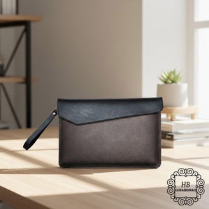 May include: A two-tone clutch with a black top flap and a brown body. The clutch has a wrist strap and a logo in the bottom right corner. The clutch is on a wooden surface.