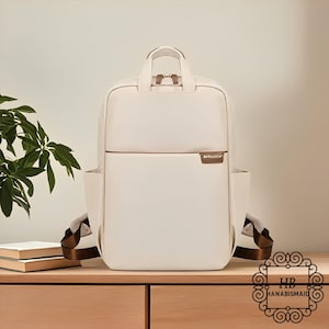 May include: Cream-colored backpack with a top handle and side pockets. The bag has brown accents and straps. The brand name "HANABISMAID" is visible in the lower right corner. The backpack is sitting on a wooden surface.