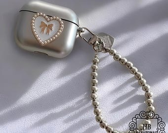 May include: Silver-colored AirPod case with a heart-shaped design. The heart features a white center, a gold bow, and a pearl border. A silver beaded chain and a clasp are attached to the case. The case has the text "HANABISMAID".