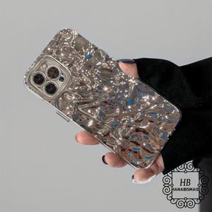 Puede incluir: Funda de teléfono plateada con una superficie texturizada y ondulada. La funda tiene un aspecto metálico reflectante y un recorte para la cámara. El logotipo de la marca "HB HANABISMAID" es visible en la esquina inferior derecha.