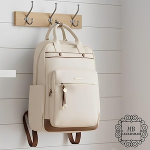May include: Cream-colored backpack with brown trim and accents, hanging on a wooden rack with metal hooks. The backpack features a front pocket with a zipper and a top handle. The bag has a rectangular shape and is designed for carrying items.