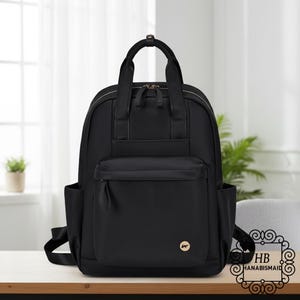 May include: A black backpack with a top handle and a front pocket. The backpack has two side pockets and adjustable shoulder straps. A small gold-colored logo is on the front. The backpack is on a wooden surface.