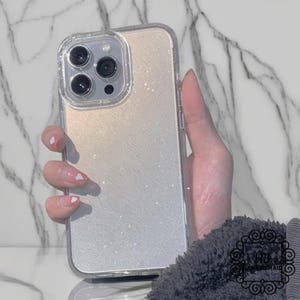 May include: Clear phone case with a glitter gradient design. The case has a clear border and a sparkly, iridescent finish that transitions from a light gold to silver. The phone case has a camera cutout with three lenses.