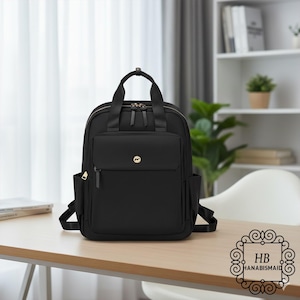 May include: A black backpack with a front pocket and side pockets, sitting on a light-colored wooden surface. The backpack has a top handle and adjustable straps. A gold-colored logo is visible on the front pocket. The brand name "HB HANABISMAID" is in the bottom right corner.