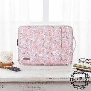 May include: A pink laptop sleeve with a floral pattern of white, gray, and peach-colored flowers. The sleeve has a zipper closure, a handle, and a small label. The sleeve is on a white wooden table.