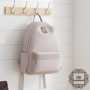 May include: A light purple backpack with gold zippers and a front pocket hangs from a wooden rack with metal hooks. The backpack has a top handle and a side pocket. The brand name "HANABISMAID" is visible.