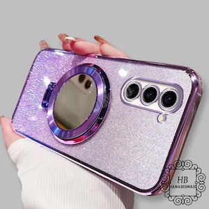 May include: A purple glitter phone case with a built-in mirror and a ring holder. The case has a clear back and a purple glitter design. The mirror is round and has a purple trim. The ring holder is silver and has a purple trim.