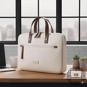 Puede incluir: Bolso para portátil de color crema con asas y detalles en cuero marrón. El bolso tiene cierre de cremallera y un pequeño logotipo rectangular. El bolso está sobre un escritorio de madera junto a un portátil, una taza y pequeñas plantas en macetas.