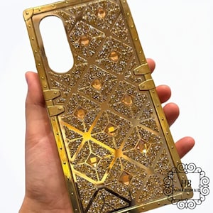 Puede incluir: Una funda de teléfono dorada con un patrón geométrico y diamantes de imitación. La funda tiene un acabado texturizado y metálico.